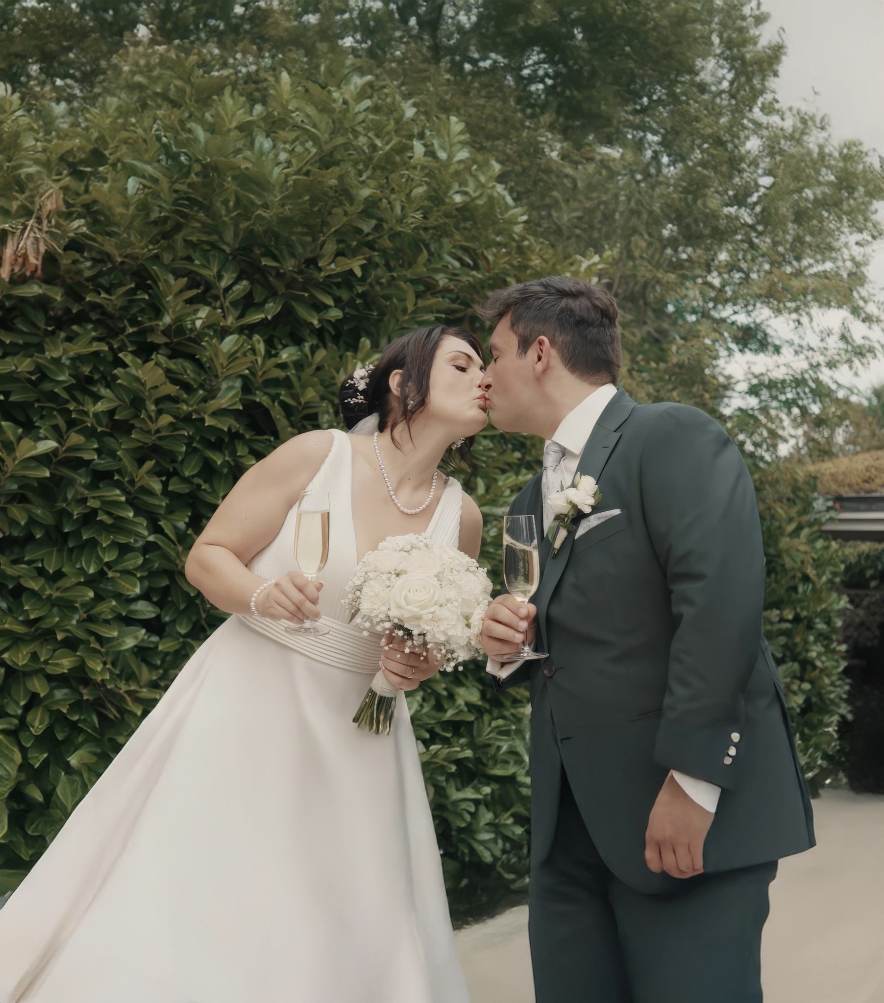 A wedding couple kissing with champagne glasses in a garden.